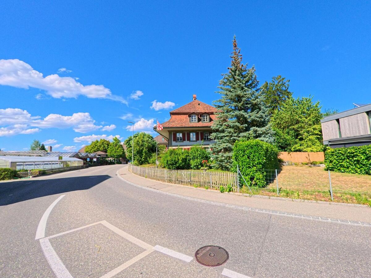 In Belp (3123, Kanton Bern) windet sich eine Dorfstrasse zwischen einem traditionellen Wohnhaus mit Schweizer Fahne, modernen Gebäuden und Gewächshäusern entlang.