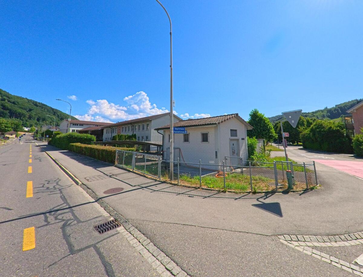 In Belp (3123, Kanton Bern) zweigt die Spitalackerstrasse von einer leicht ansteigenden Hauptstrasse ab, flankiert von Dienstgebäuden und einem markierten Radweg unter sommerlicher Sonne.