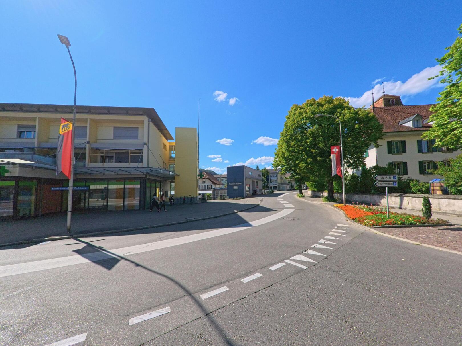 In Belp (3123, Kanton Bern) zweigt eine innerörtliche Strasse an einem blühenden Kreisbeet und einem historischen Gebäude mit Schweizer Flagge vom modernen Zentrum mit Apotheke ab.