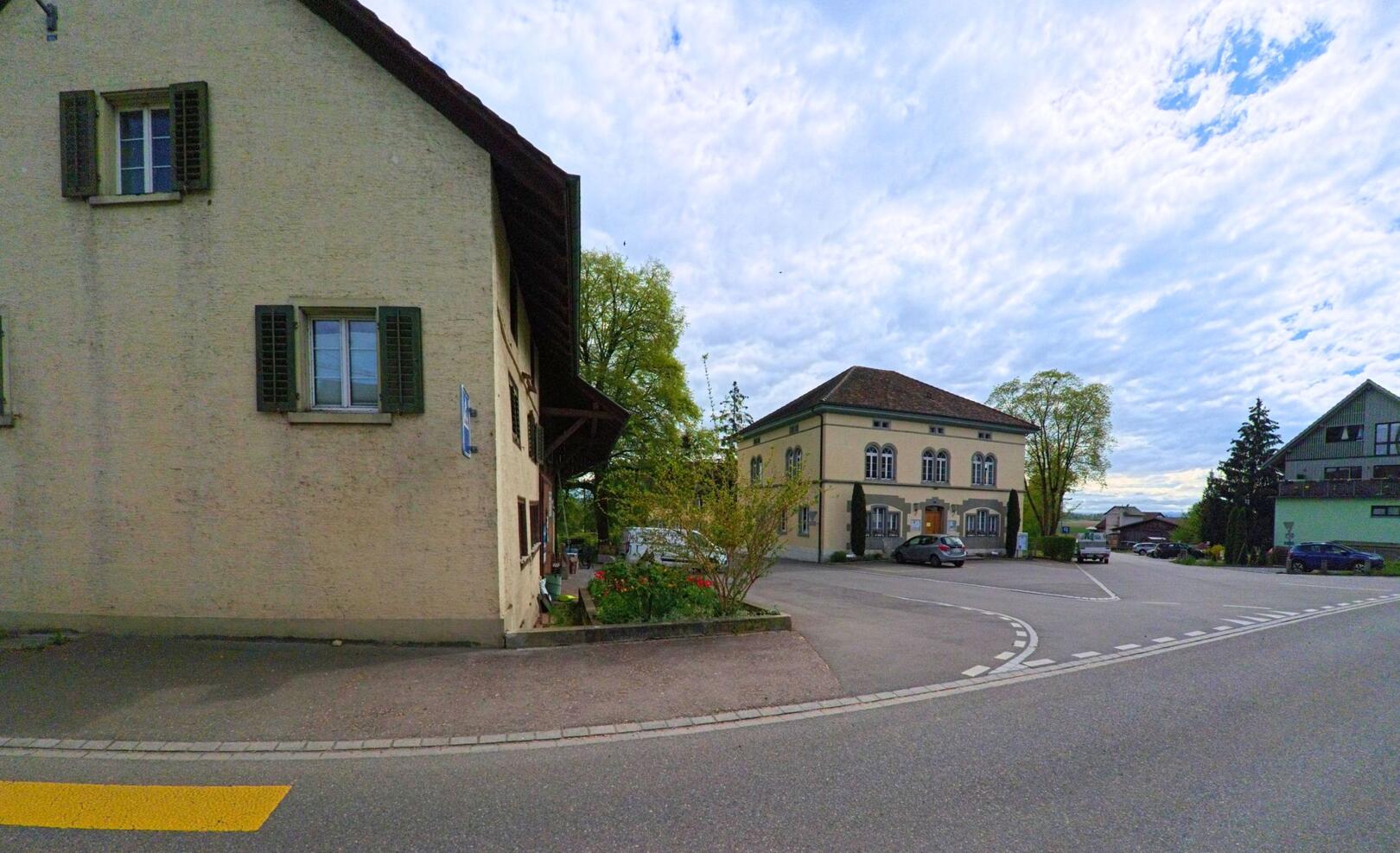 In Benken (8463, Kanton Zürich) führt die Strasse an einem historischen Gebäude vorbei, mit einer grossen Wiese und einer ruhigen Umgebung im Hintergrund.