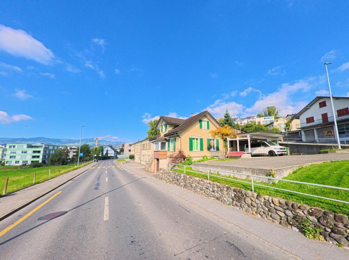 Eine leicht ansteigende Strasse mit Wohnhaeusern und offenen Gruenflaechen fuehrt durch Benken (8717, Kanton St. Gallen) bei sonnigem Himmel.