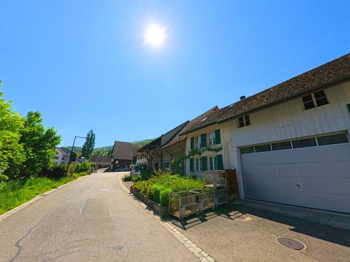 In Bennwil (4431, Kanton Baselland) verläuft eine ruhige Strasse an einem traditionellen Wohnhaus mit grünem Fensterladen und Vorgarten vorbei in Richtung Dorfkern.