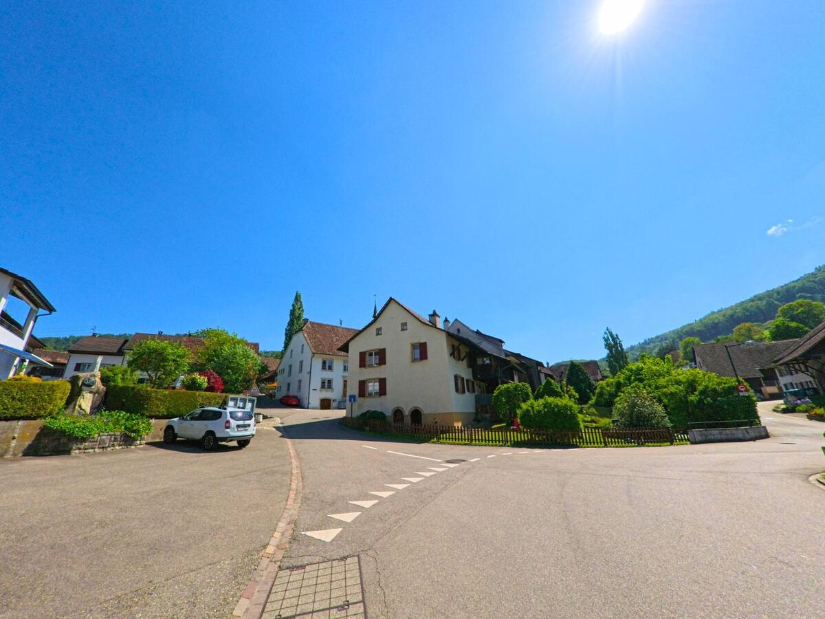 In Bennwil (4431, Kanton Baselland) mündet eine Wohnstrasse in einen kleinen Platz mit gepflegten Vorgärten und traditionellen Häusern vor bewaldeter Hangkulisse.