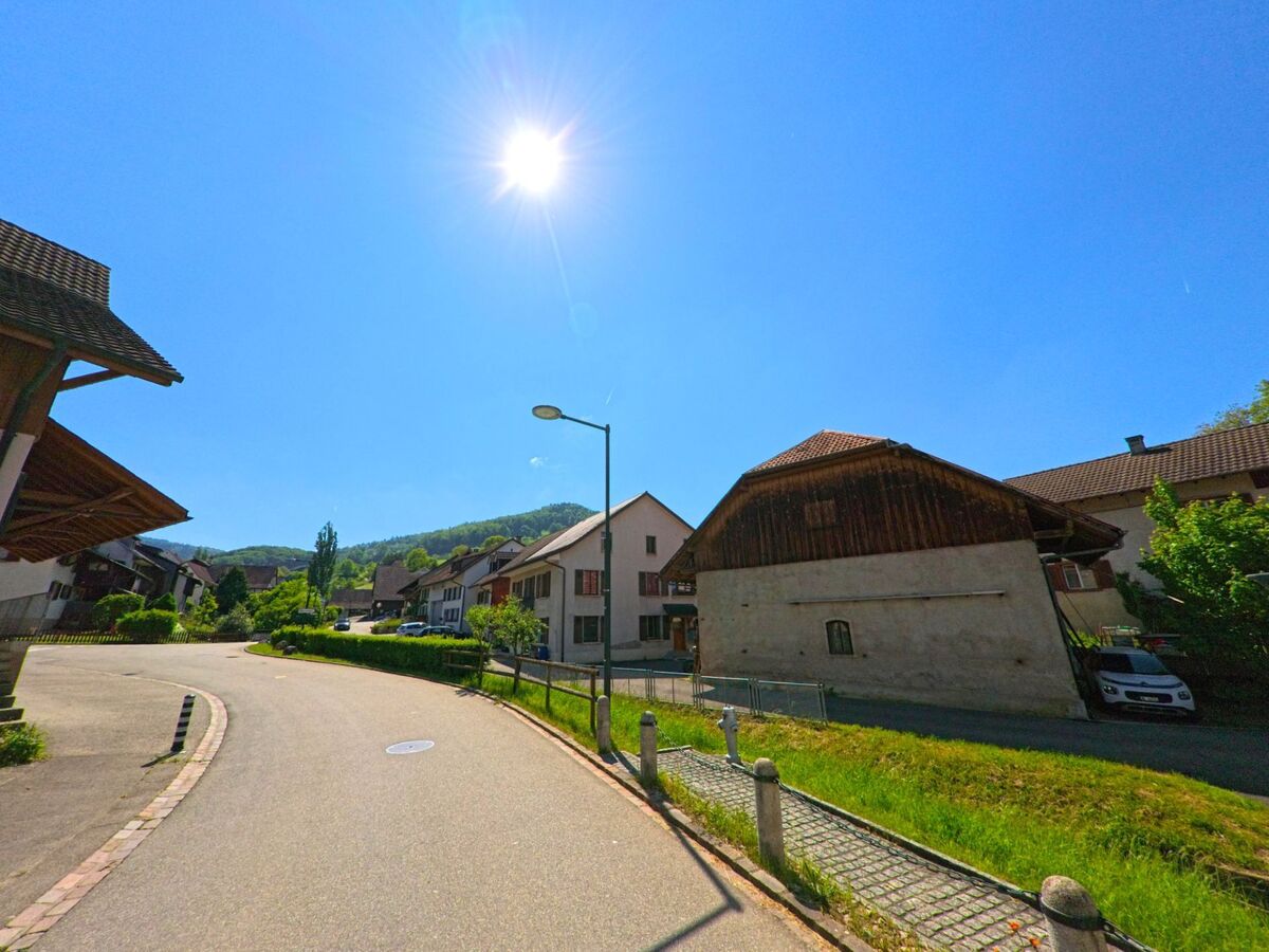 In Bennwil (4431, Kanton Baselland) schlängelt sich eine Wohnstrasse bei Sonnenschein durch das Dorf, vorbei an Scheunen und Einfamilienhäusern mit Blick auf bewaldete Hügel.