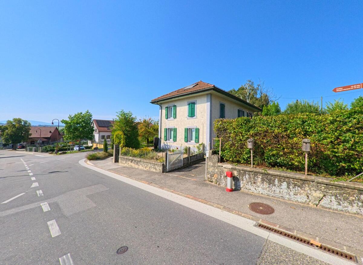 Ein traditionelles Wohnhaus mit grünen Fensterlaeden steht in Bercher (1038, Kanton Waadt) an einer Strasse mit Wegweisern und angrenzender Hecke.