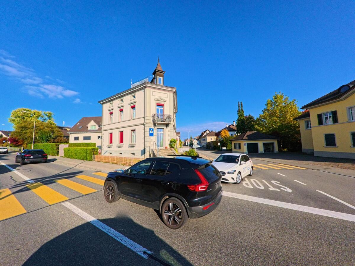 An einer gut markierten Kreuzung mit Fussgängerstreifen steht ein repräsentatives Gebäude im Ortskern von Berg (8572, Kanton Thurgau) unter klarem blauem Himmel.