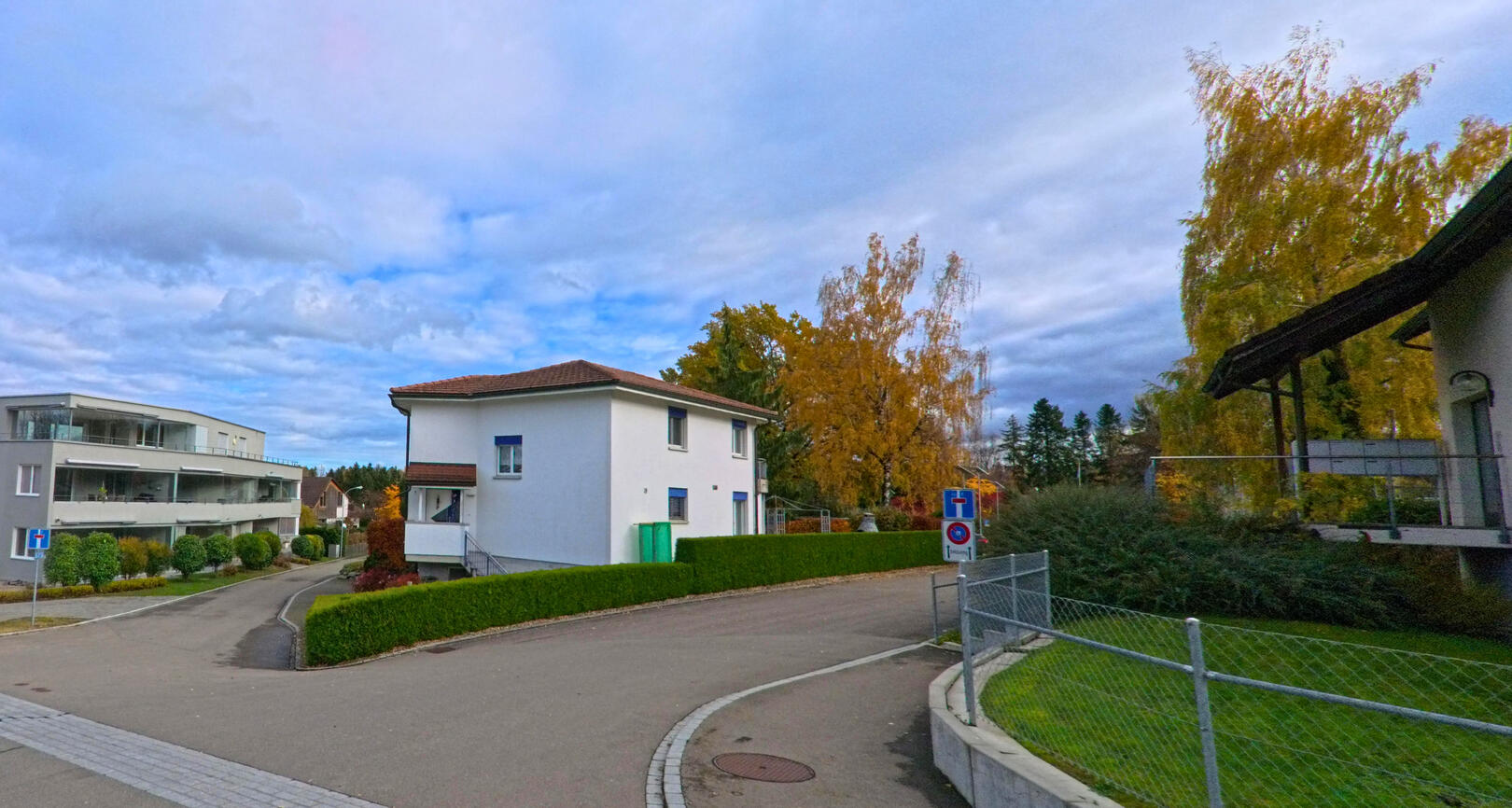 In Berg (9305, Kanton St. Gallen) erstreckt sich eine ruhige Wohnstrasse, flankiert von modernen Häusern und herbstlich gefärbten Bäumen, die den Himmel überragen.