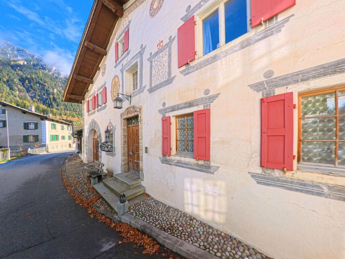 Entlang einer schmalen Dorfstrasse zeigt sich ein historisches Engadiner Haus mit bemalter Fassade und roten Fensterläden in Bergün_Filisur (7477, Kanton Graubünden).