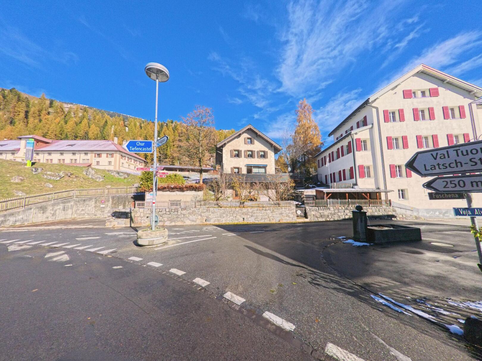 An einer zentralen Wegkreuzung mit Hinweistafeln stehen Wohn- und Verwaltungsgebäude in Bergün_Filisur (7482, Kanton Graubünden), umgeben von herbstlichen Bäumen und Hanglagen.