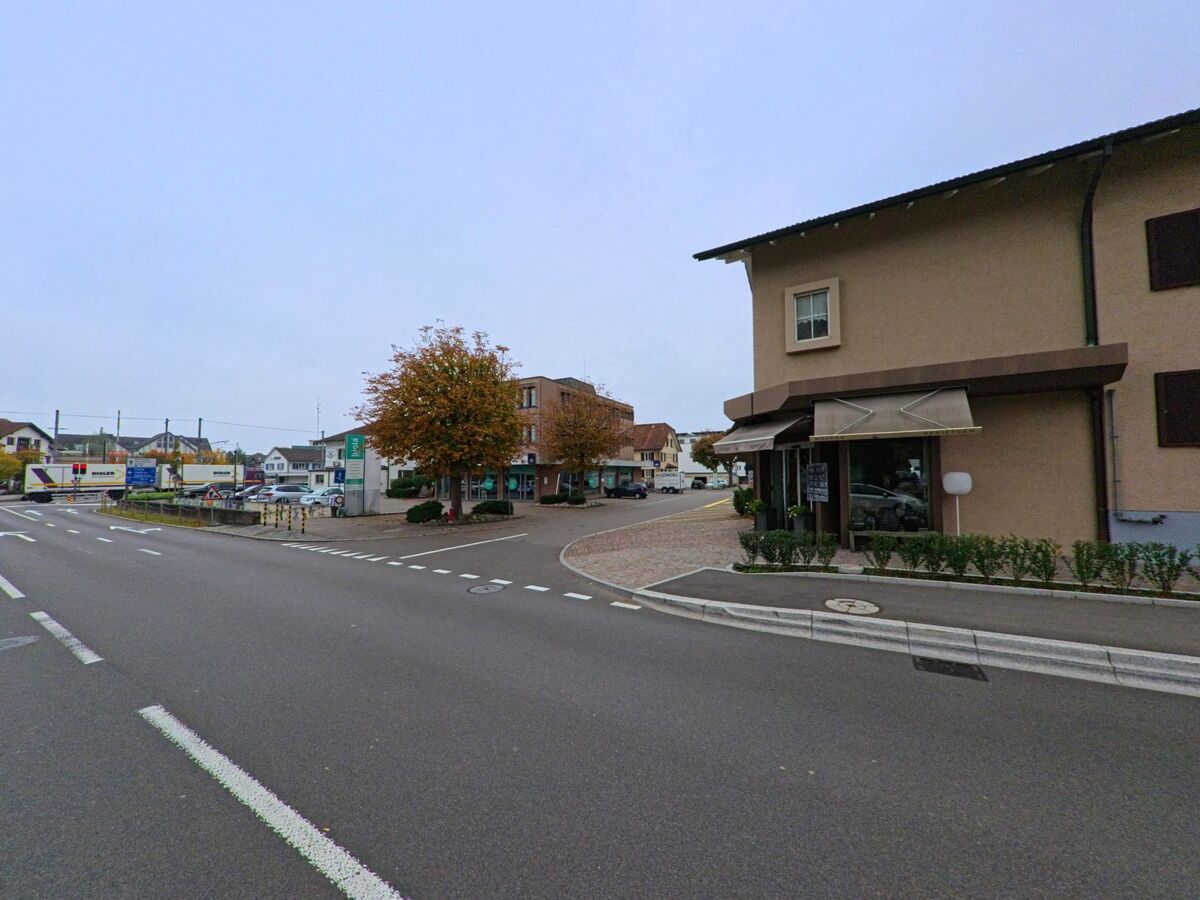 Eine Kreuzung in Berikon (8965, Kanton Aargau) mit Geschäften, Bäumen und sichtbaren Bahngeleisen im Hintergrund vermittelt ein belebtes Ortszentrum.