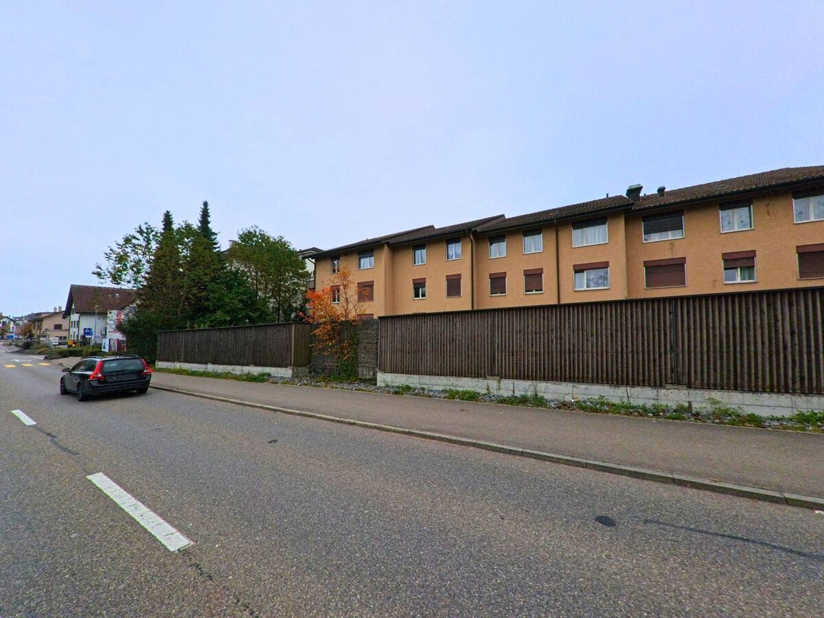 Entlang einer Hauptstrasse in Berikon (8965, Kanton Aargau) steht ein langgezogenes Wohnhaus mit brauner Fassade und geschlossenen Fensterläden hinter einem Holzzaun.