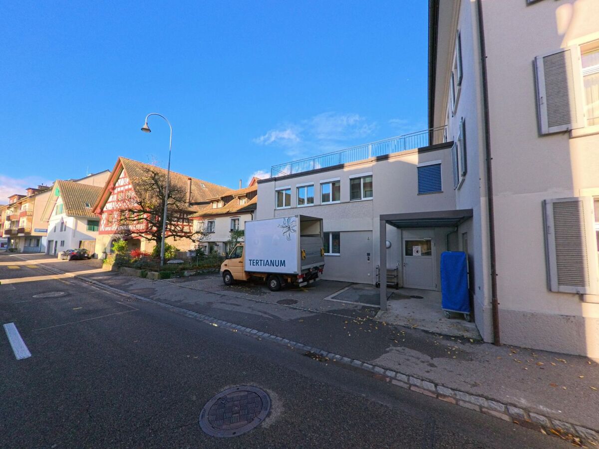Vor einem zeitgemässen Gebäude mit Anlieferungsbereich verläuft eine ruhige Strasse in Berlingen (8267, Kanton Thurgau), während im Hintergrund historische Häuser das Ortsbild ergänzen.