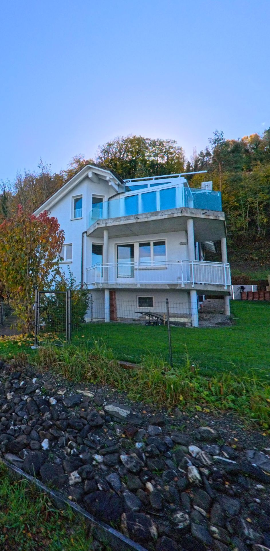 Am Hang oberhalb eines grünen Gartens steht ein Wohnhaus mit Balkon und Glasbrüstung in Berlingen (8267, Kanton Thurgau), umgeben von herbstlicher Vegetation.