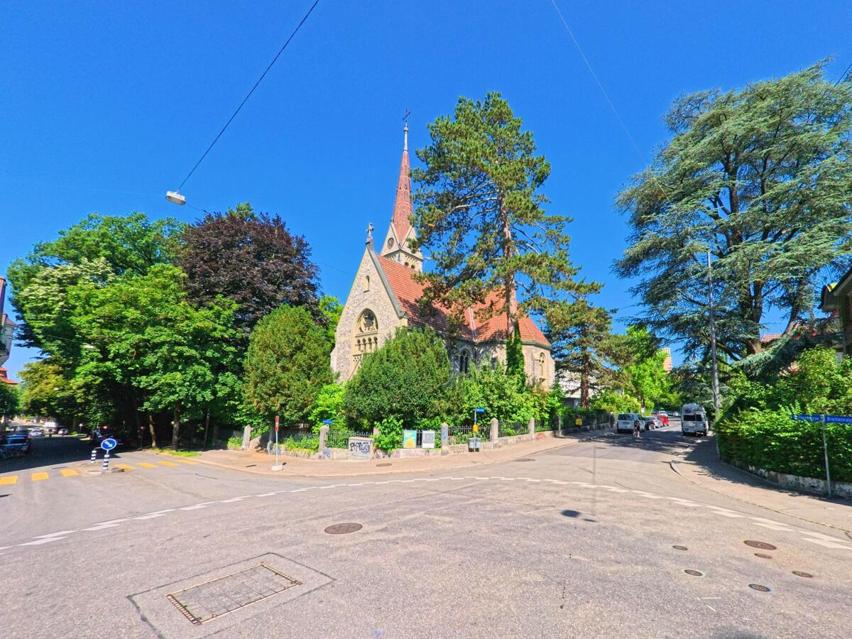 In Bern (3014, Kanton Bern) liegt eine Kirche mit spitzem Turm eingebettet zwischen hohen Bäumen an einer ruhigen Kreuzung mit Zebrastreifen und Hinweisschildern.