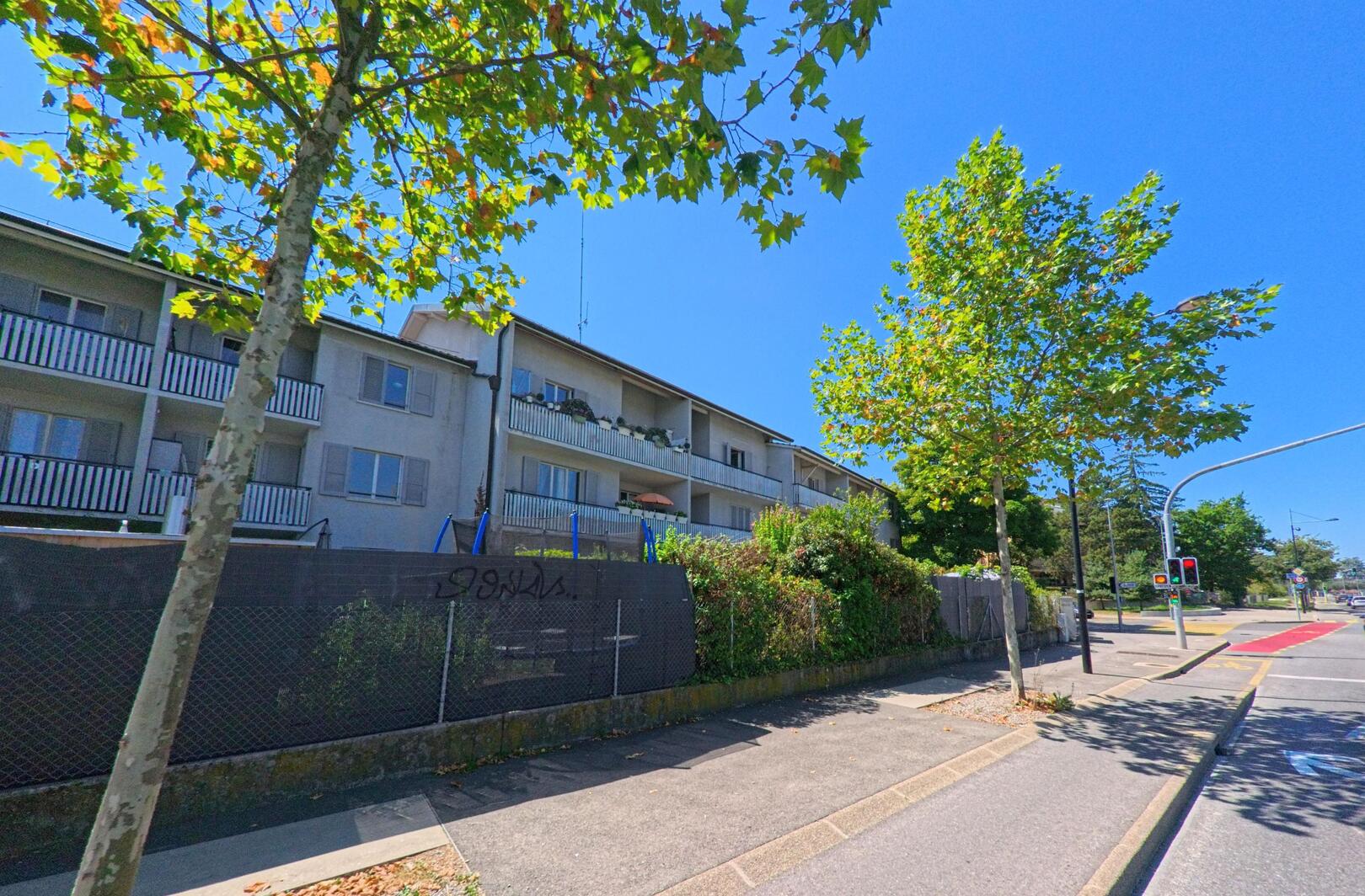 In Bernex (1233, Kanton Genf) stehen mehrstöckige Wohnhäuser mit Balkonen hinter einer Strasse mit Ampelanlage und jungen Bäumen.