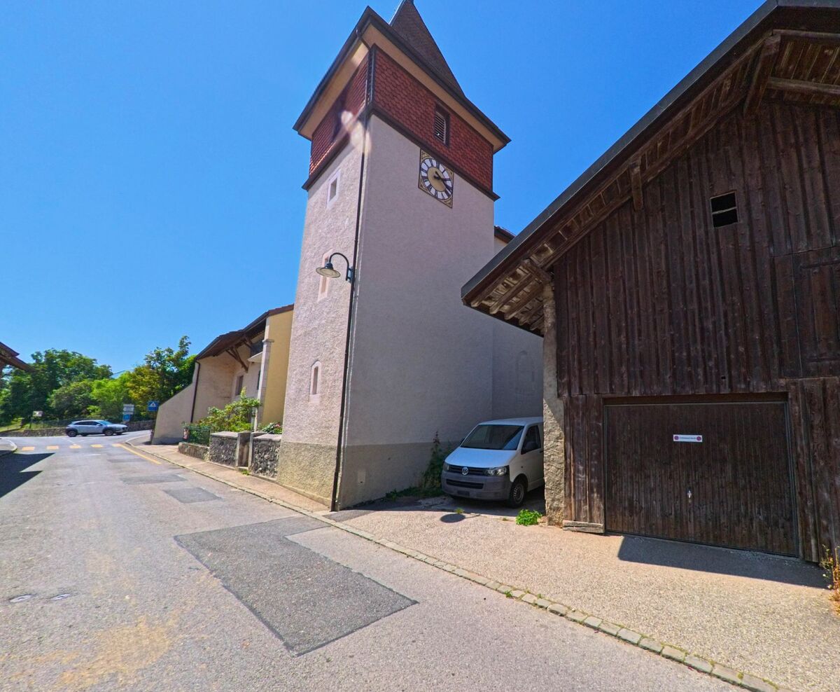 Der Kirchturm von Berolle (1149, Kanton Waadt) mit Uhr ragt neben einem Holzgebäude an einer schmalen Strasse auf.