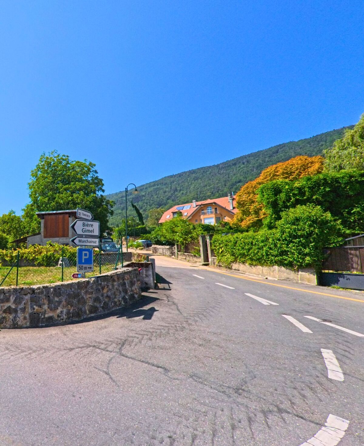 Ein Strassenschild in Berolle (1149, Kanton Waadt) weist den Weg nach Biere, Gimel und Marchairuz, im Hintergrund erhebt sich der bewaldete Hügel.