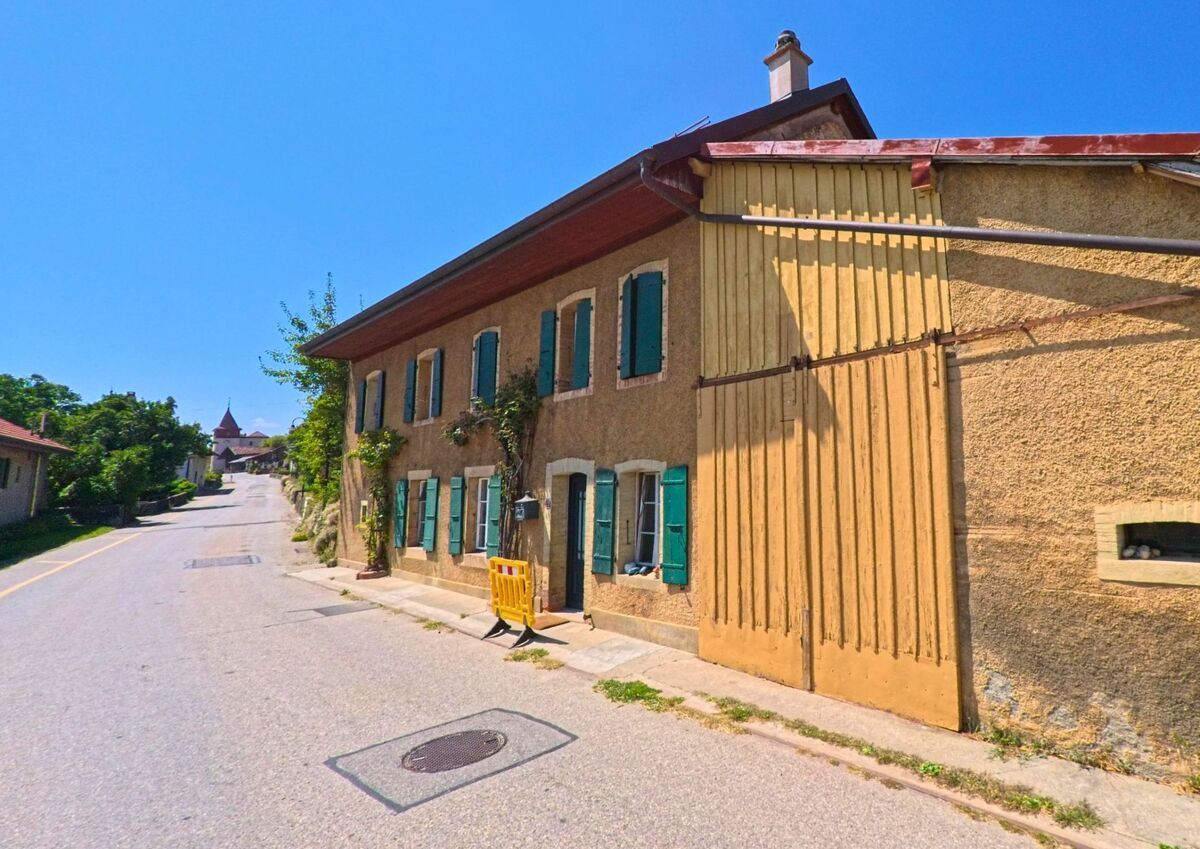 Ein traditionelles Wohnhaus mit grünen Fensterläden und angebauter Scheune steht an einer Strasse in Berolle (1149, Kanton Waadt).