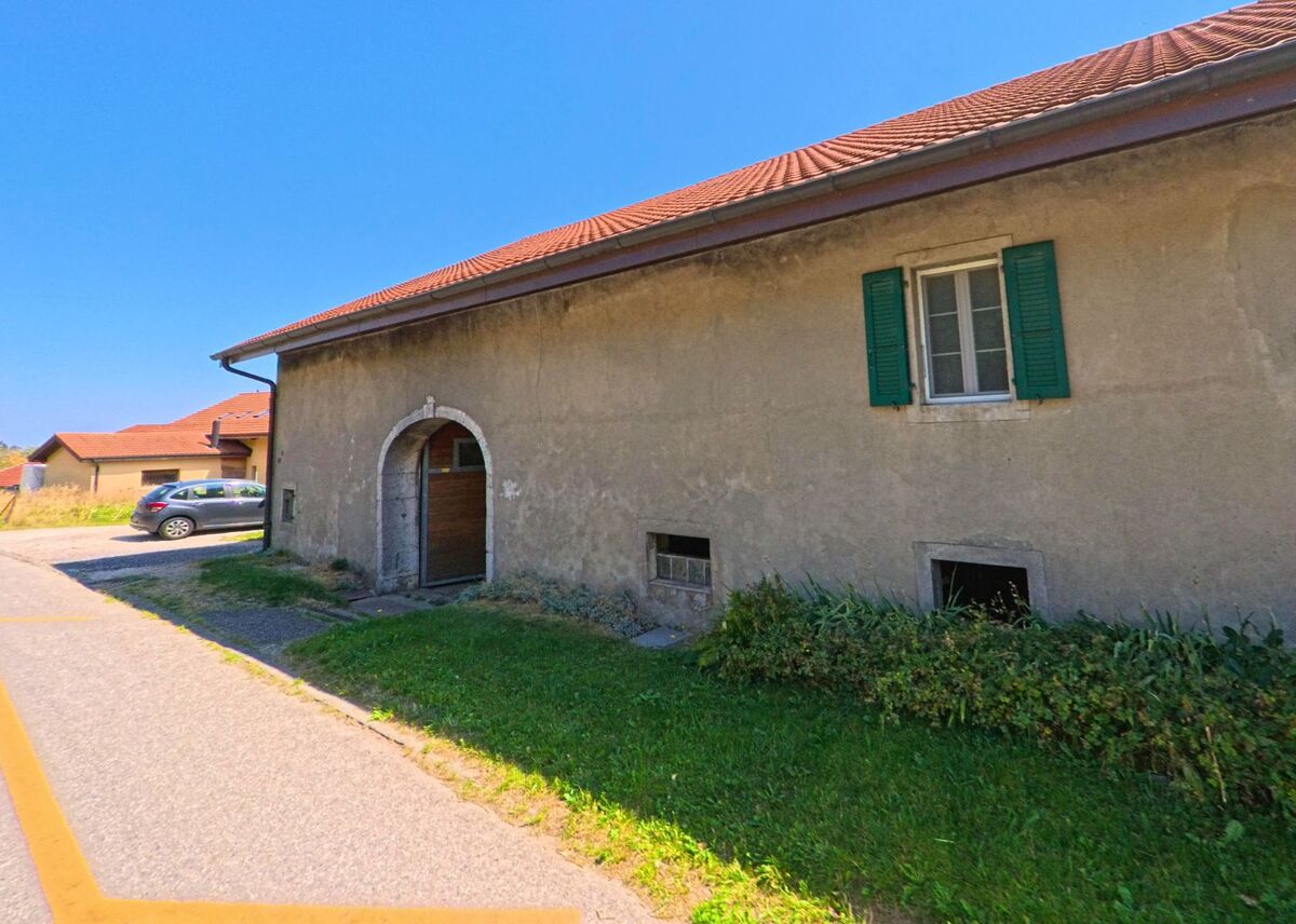 Ein älteres Wohnhaus in Berolle (1149, Kanton Waadt) zeigt eine einfache Fassade mit gruenen Fensterläden und einem Torbogen.