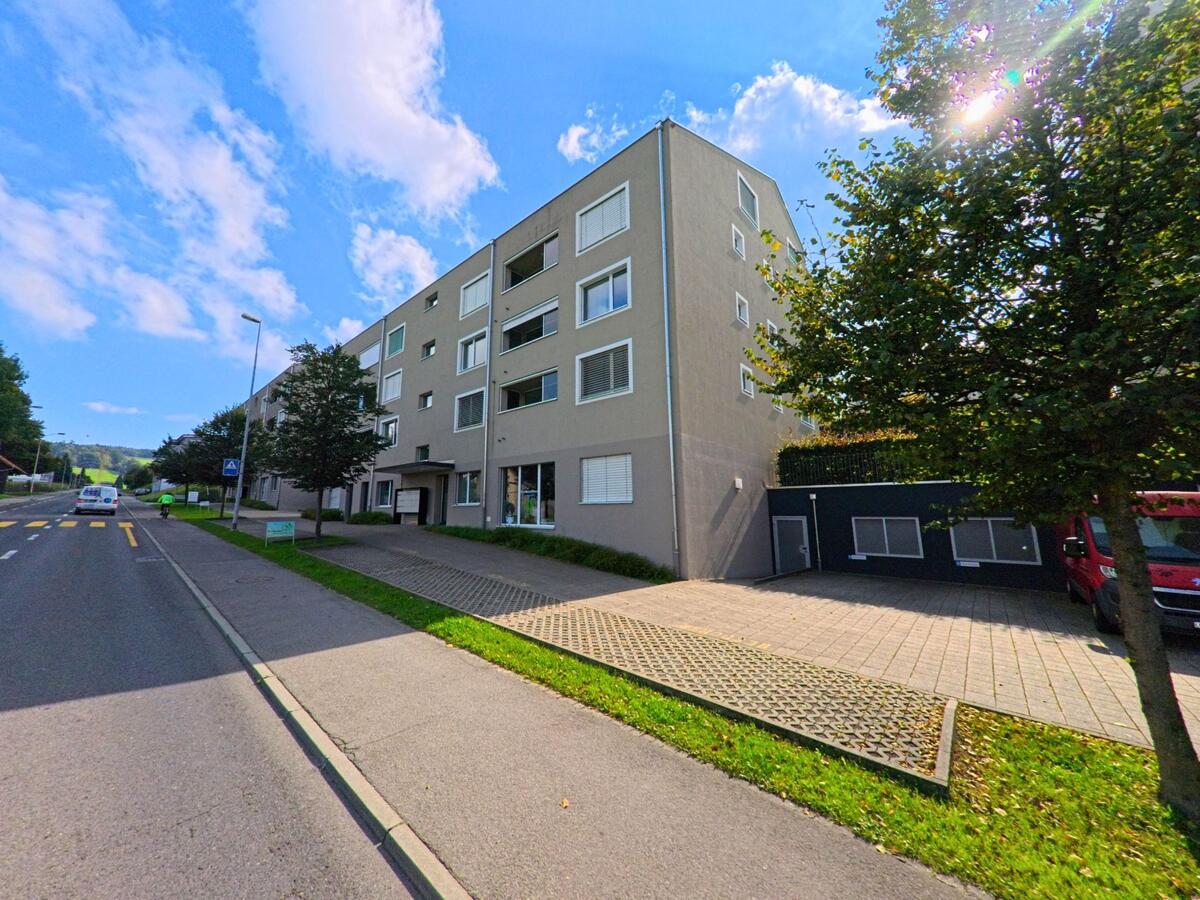 In Beromuenster (6215, Kanton Luzern) saeumt ein langgestrecktes Wohnhaus mit grossen Fenstern die Strasse, die an einem Zebrastreifen und jungen Baumen vorbeifuehrt.