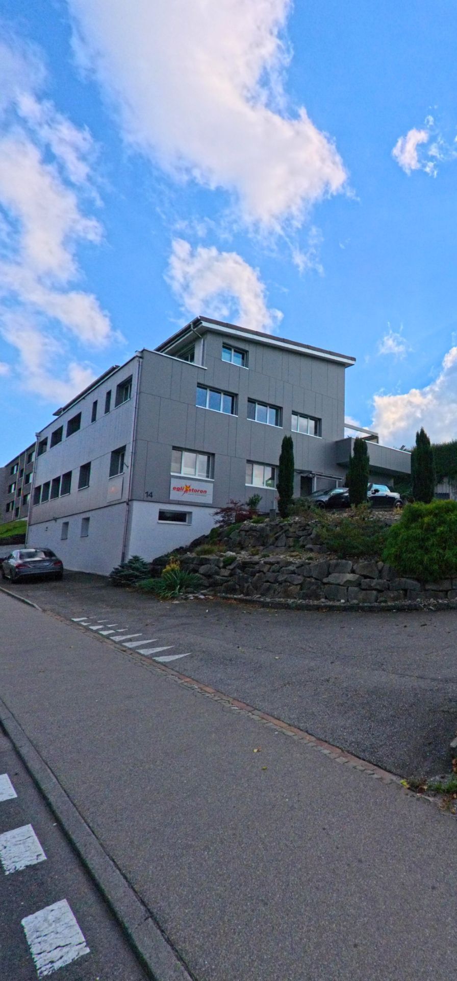 In Beromuenster (6215, Kanton Luzern) steht ein modernes, graues Gebaeude an einer leicht ansteigenden Strasse, flankiert von einer bepflanzten Steinmauer.