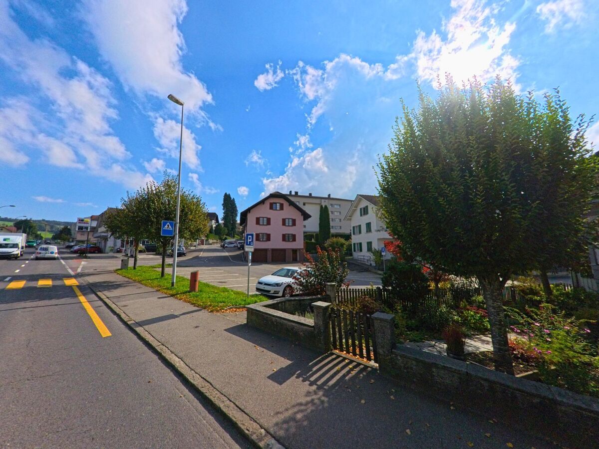 In Beromuenster (6215, Kanton Luzern) verlaeuft eine breite Strasse mit Zebrastreifen an Wohnhaeusern und kleinen Gaerten vorbei, waehrend im Hintergrund ein Parkplatz und weitere Gebaeude sichtbar sind.