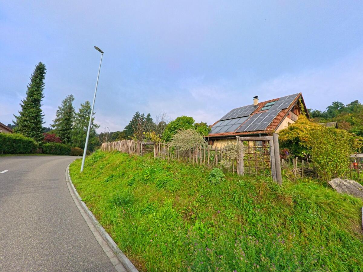Ein Einfamilienhaus mit Solarpanels auf dem Dach steht in Besenbüren (5627, Kanton Aargau) an einer leicht kurvigen Strasse mit Grünstreifen und Holzzaun.