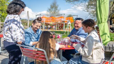 Im Besucherzentrum Kinderdorf Pestalozzi in Trogen (9043, Kanton Appenzell Ausserrhoden) sitzt eine Familie bei sonnigem Wetter an einem roten Tisch im Gartenbereich und geniesst die gemeinsame Zeit.