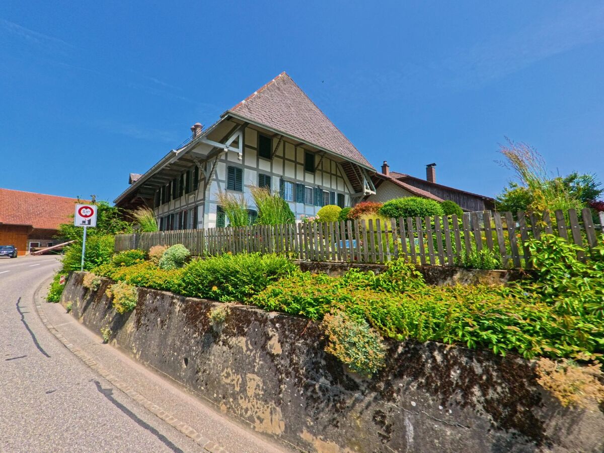 Ein grosses Bauernhaus mit gepflegtem Garten und typischem Giebeldach erhebt sich in Bettenhausen (3366, Kanton Bern) über eine leicht ansteigende Strasse mit Verkehrsschild.