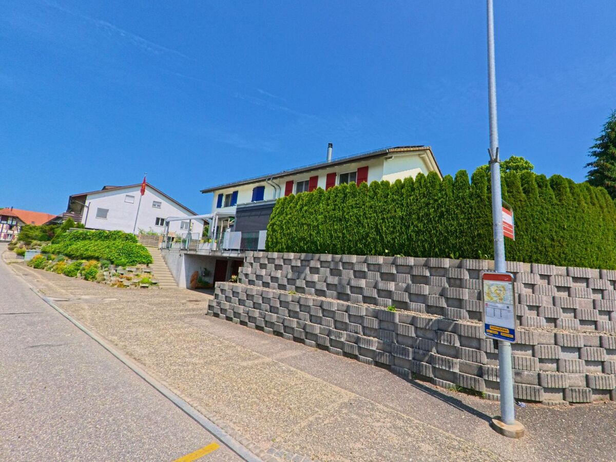 In Bettenhausen (3366, Kanton Bern) verläuft eine Strasse mit Bushaltestelle entlang einer erhöht liegenden Häuserzeile mit sauber gestalteten Stützmauern und Hecken.