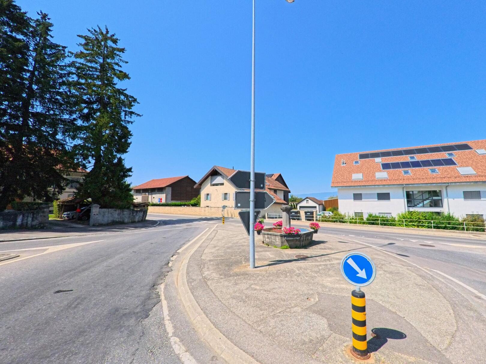 Eine Verkehrsinsel mit Blumenschmuck und Strassenschildern befindet sich in Bettens (1042, Kanton Waadt) zwischen Wohnhaeusern und einem Gebaeude mit Solarpanelen.