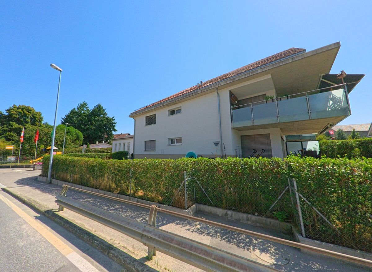 Ein modernes Wohnhaus mit grossem Balkon und gepflegter Hecke steht in Bettens (1377, Kanton Waadt) an einer Strasse mit Strassenlaterne.