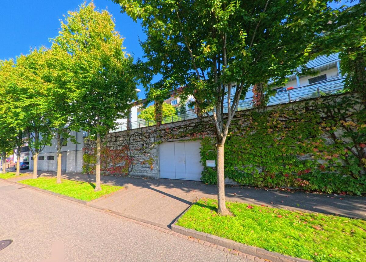 Entlang eines Gehwegs mit Bäumen und einer begrünten Stützmauer verläuft eine ruhige Strasse in Bettingen (4126, Kanton Basel-Stadt) bei klarem Wetter.