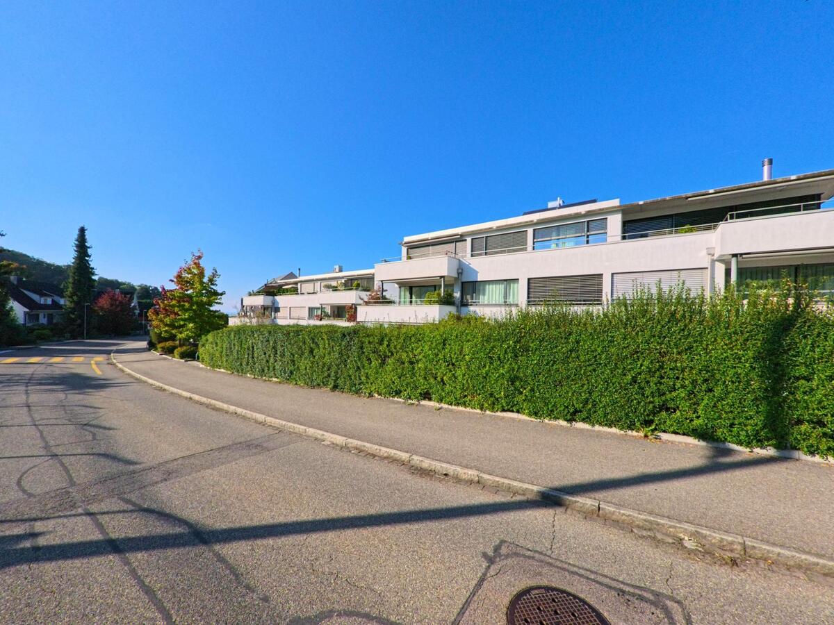 In Bettingen (4126, Kanton Basel-Stadt) saeumen moderne, weiss verputzte Mehrfamilienhaeuser und dichte Hecken eine sanft geschwungene Wohnstrasse bei klarem Himmel.
