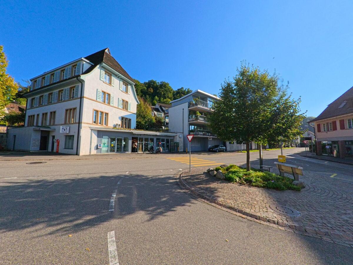 Vor einem kleinen Platz mit Rundbeet stehen in Bettingen (4126, Kanton Basel-Stadt) ein traditionelles Mehrstockhaus und moderne Wohnbauten unter klarem Himmel.