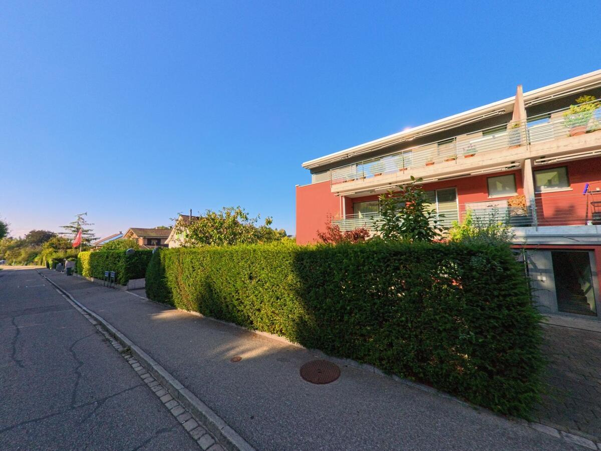In Bettingen (4126, Kanton Basel-Stadt) verläuft eine ruhige Wohnstrasse entlang dichter Hecken und eines modernen Hauses mit roter Fassade und Balkonen.