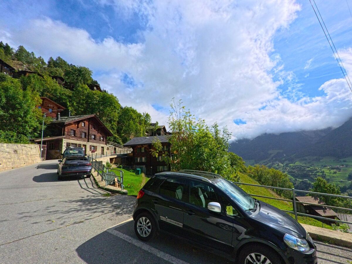 Am Hang von Bettmeralp (3991, Kanton Wallis) stehen Holzhäuser und geparkte Autos mit Blick ins grüne Tal.