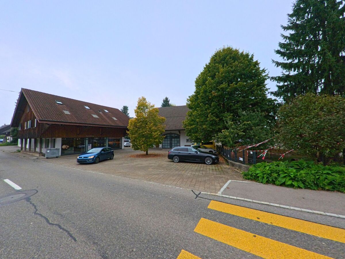 Ein grosses Haus mit Holzfassade und Satteldach steht an einer Strasse in Bettwil (5618, Kanton Aargau), daneben befinden sich Parkplaetze und Baeume.