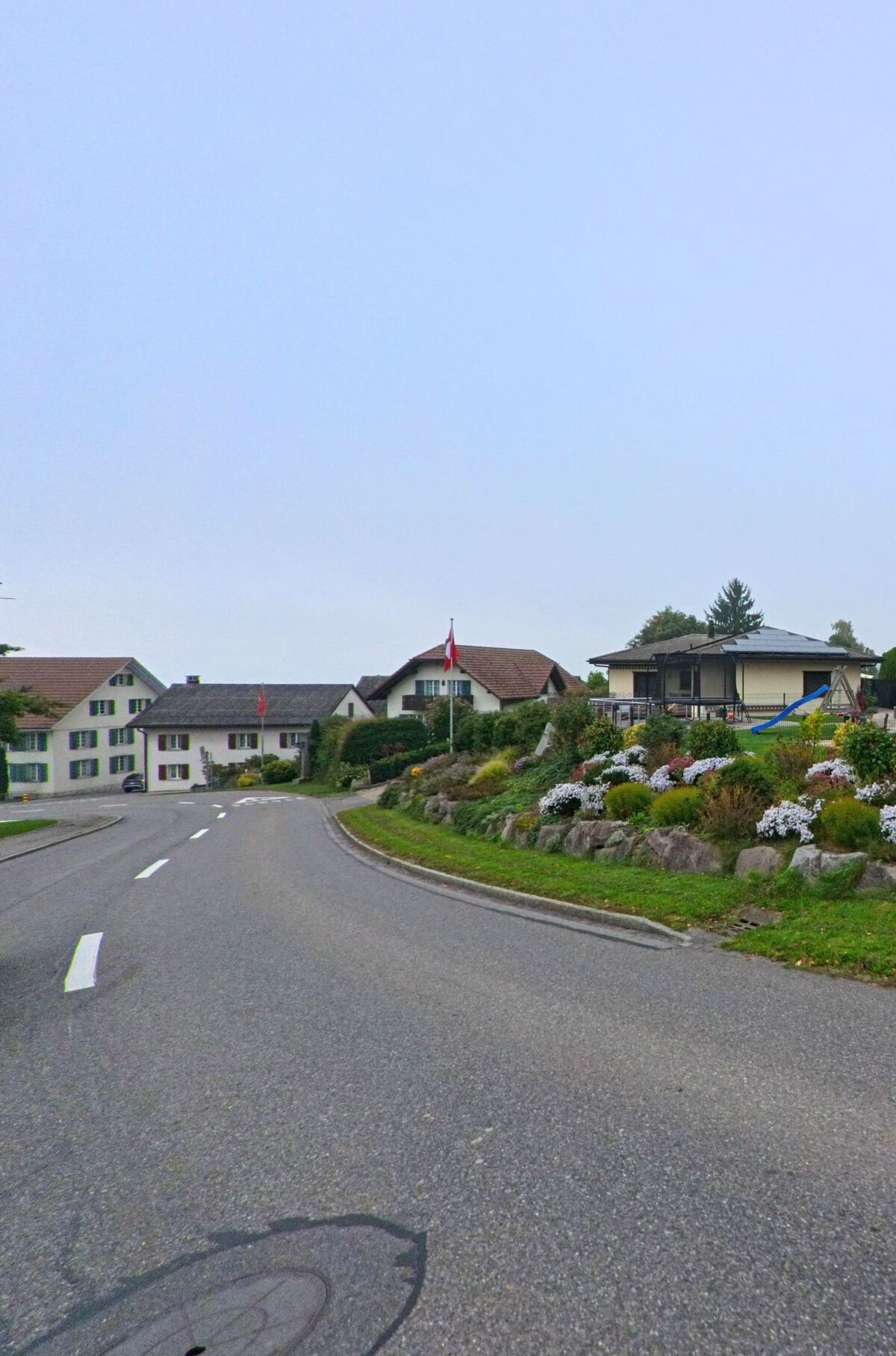 An einer leicht gekruemmten Strasse in Bettwil (5618, Kanton Aargau) saeumen gepflegte Haeuser und ein bepflanzter Kreisel mit Schweizer Fahne das Dorfbild.