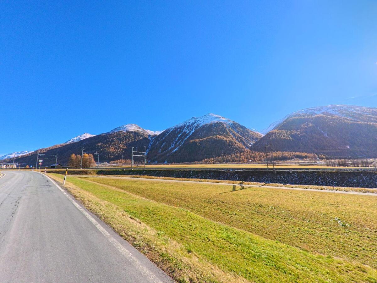 Entlang einer ruhigen Landstrasse in Bever (7502, Kanton Graubünden) erstrecken sich offene Wiesenflächen vor einem breiten Bergrücken mit schneebedeckten Gipfeln.