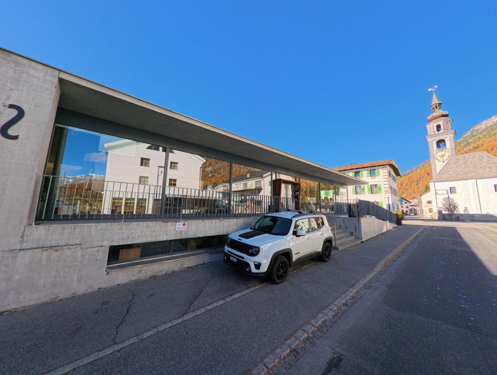 An einer ruhigen Dorfstrasse in Bever (7502, Kanton Graubünden) steht ein modernes Gebäude mit grosser Glasfront, während im Hintergrund die Kirche das Ortsbild prägt.