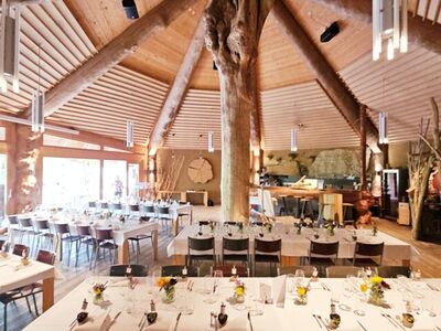 Die Biberburg in Hirschthal (5042, Kanton Aargau) bietet einen grosszügigen Gastraum mit Holzdachkonstruktion um einen zentralen Baumstamm und festlich gedeckten Tischen.