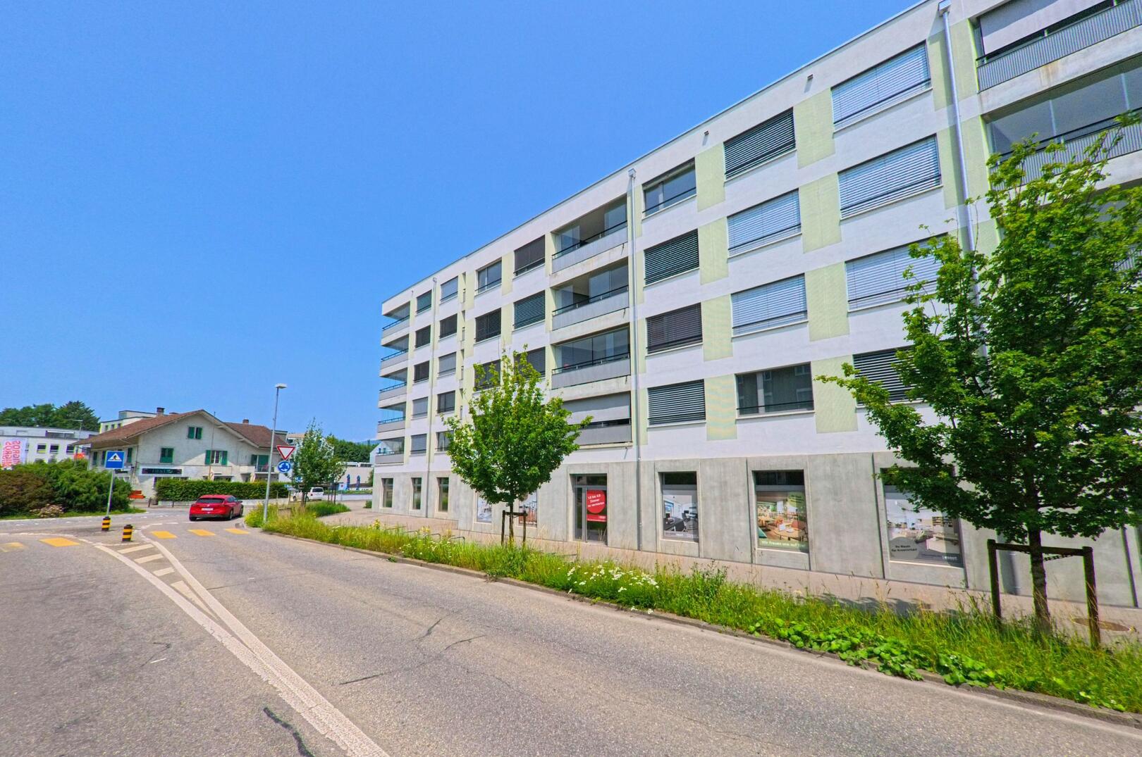 In Biberist (4562, Kanton Solothurn) steht ein modernes mehrstöckiges Wohn- und Geschäftshaus an einer Kreuzung mit Zebrastreifen und Verkehrsinsel.