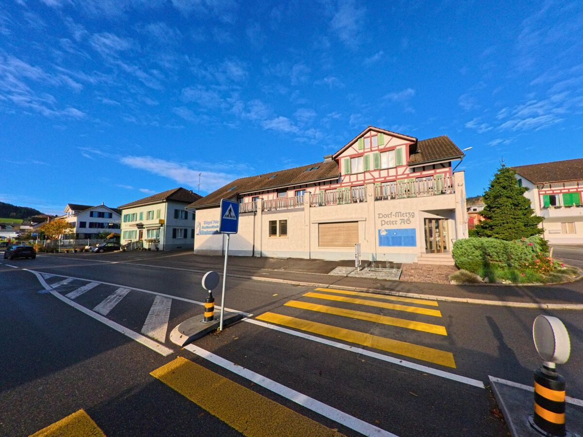 An einer Kreuzung in Bichelsee-Balterswil (8362, Kanton Thurgau) steht ein markantes Gebäude mit einem Laden und einer grossen Eingangstür neben einem Fussgängerüberweg und weiteren Wohnhäusern.