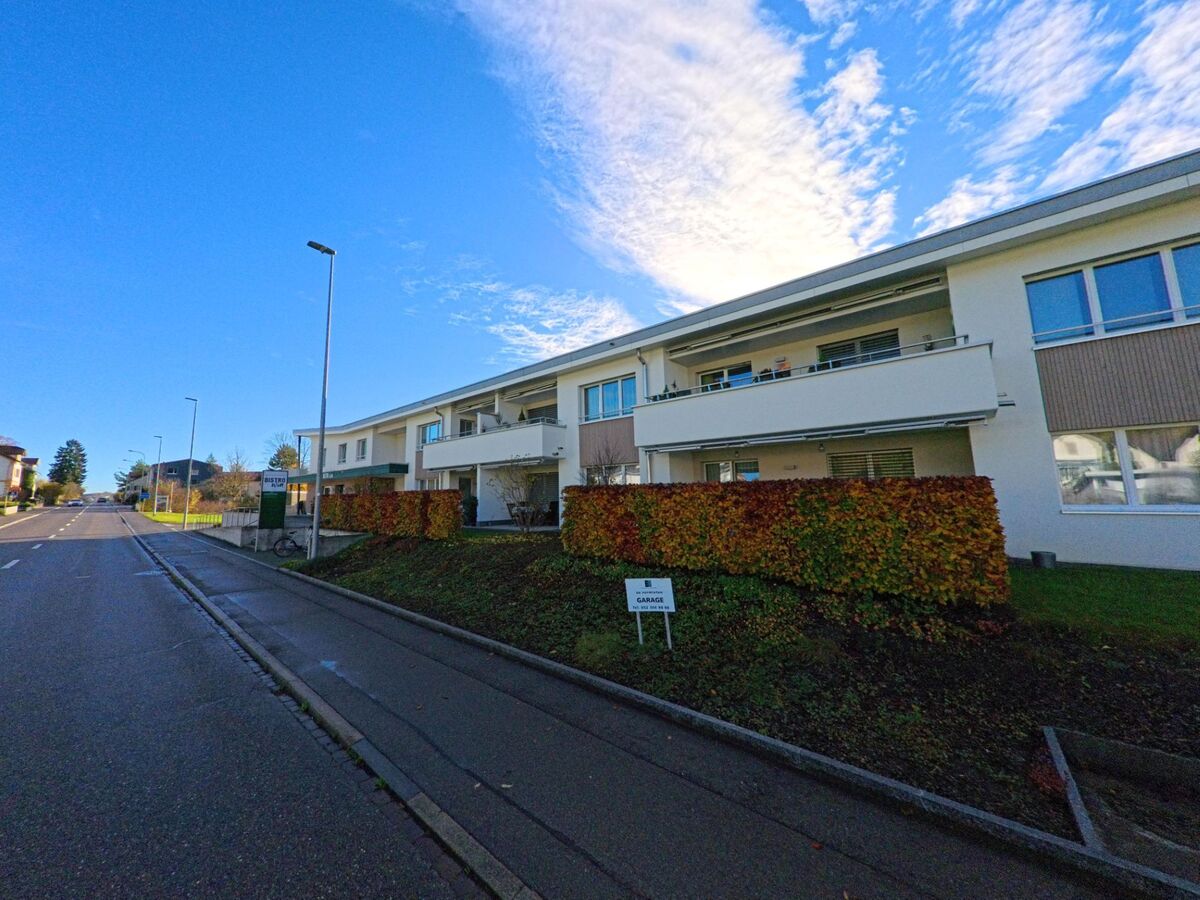 Entlang einer ruhigen Strasse in Bichelsee-Balterswil (8362, Kanton Thurgau) erstreckt sich ein modernes Mehrfamilienhaus mit Balkonen, Hecken und Gehweg unter hellem Himmel.