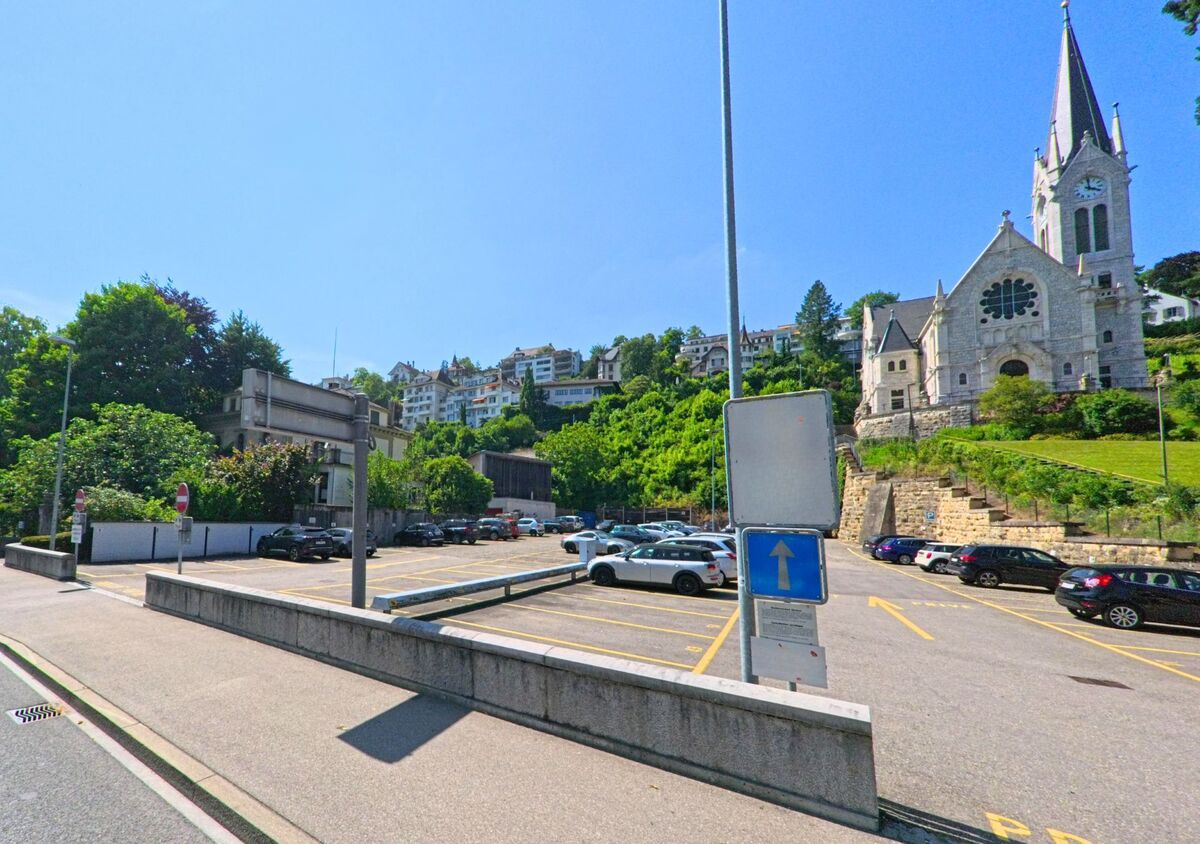 In Biel/Bienne (2500, Kanton Bern) führt die Strasse vorbei an einem privaten Parkplatz und einer Kirche, die sich majestätisch auf einem Hügel erhebt, umgeben von grünen Bäumen.