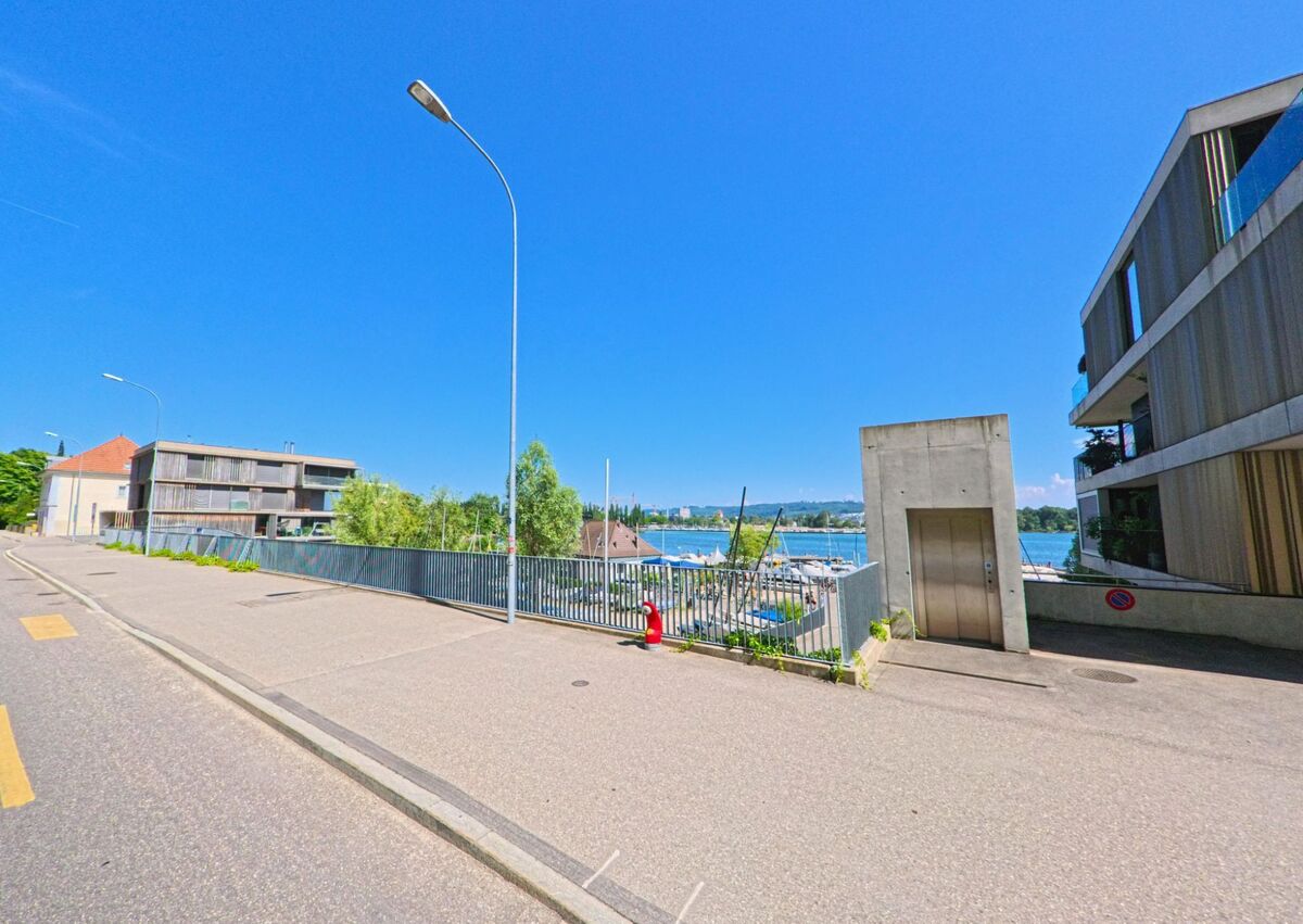 In Biel/Bienne (2500, Kanton Bern) führt die Strasse entlang moderner Wohngebäude mit Blick auf den See und die Yachten, umgeben von klar blauem Himmel.