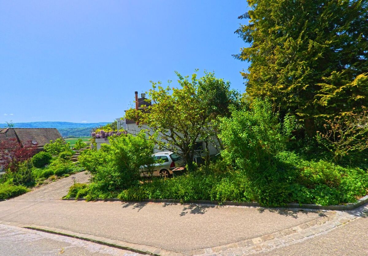In Biel-Benken (4105, Kanton Baselland) befindet sich eine begrünte Verkehrsinsel zwischen Wohnstraßen, umgeben von Bäumen, Sträuchern und Einfamilienhäusern mit Blick auf die Hügel.