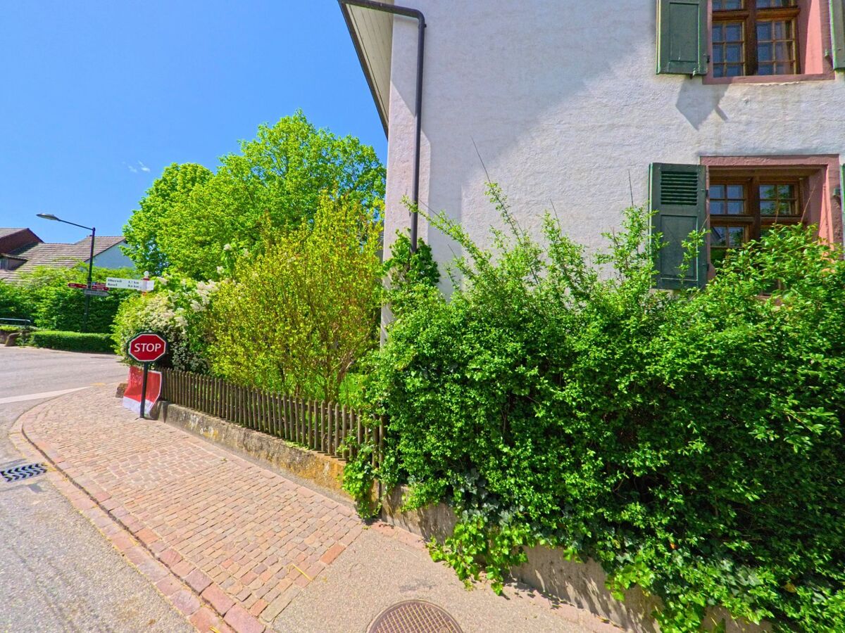 In Biel-Benken (4105, Kanton Baselland) steht ein historisches Gebäude mit Fensterläden an einer gepflasterten Straßenecke, die von einem kleinen Stoppschild und dichter Vegetation gesäumt wird.