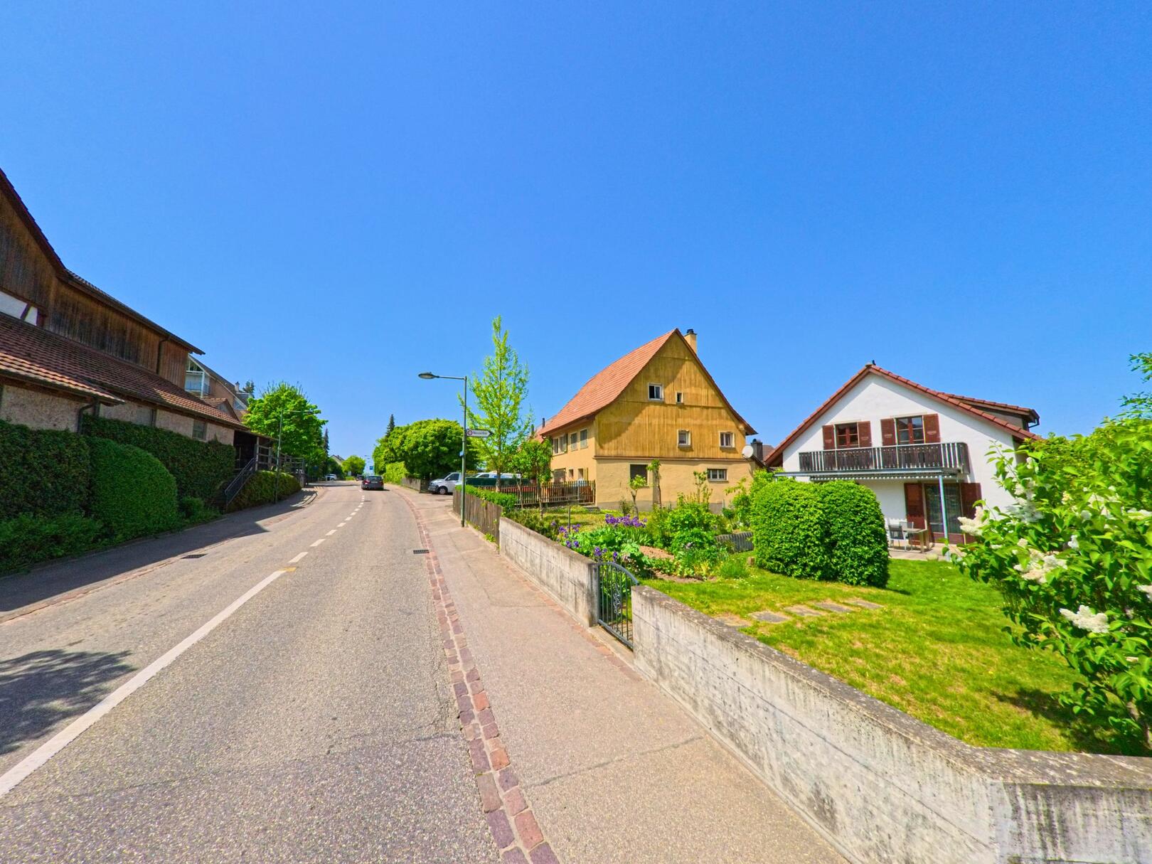 In Biel-Benken (4105, Kanton Baselland) verläuft eine leicht ansteigende Strasse entlang gepflegter Wohnhäuser und traditioneller Gebäude mit Gärten.